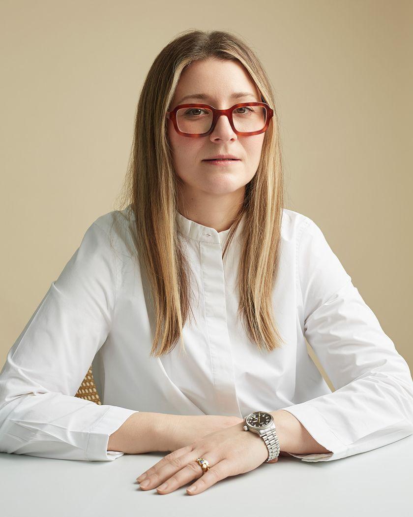 A three quarter portrait of a blond woman with large, wide brown glasses, in a mock neck white blouse
