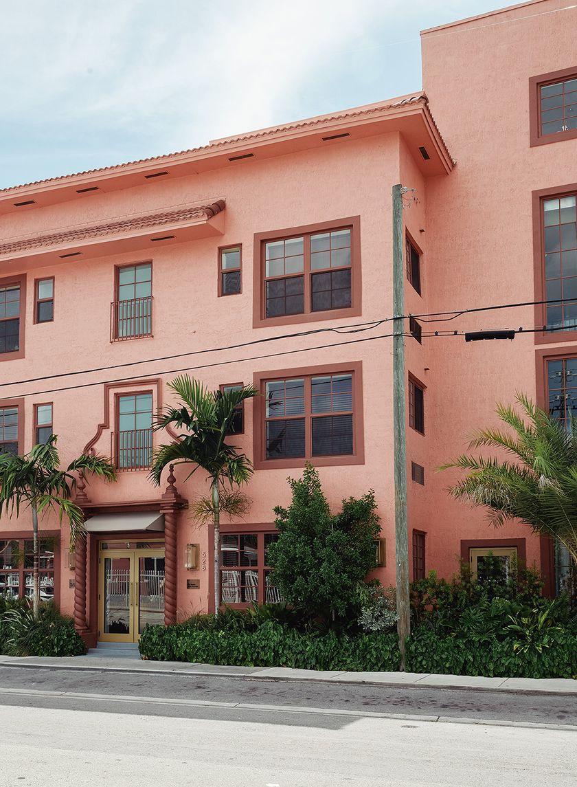 The exterior of a salmon color Cuban-colonial style building with burgundy window frames and luscious palm trees framing the brass door and entrance area