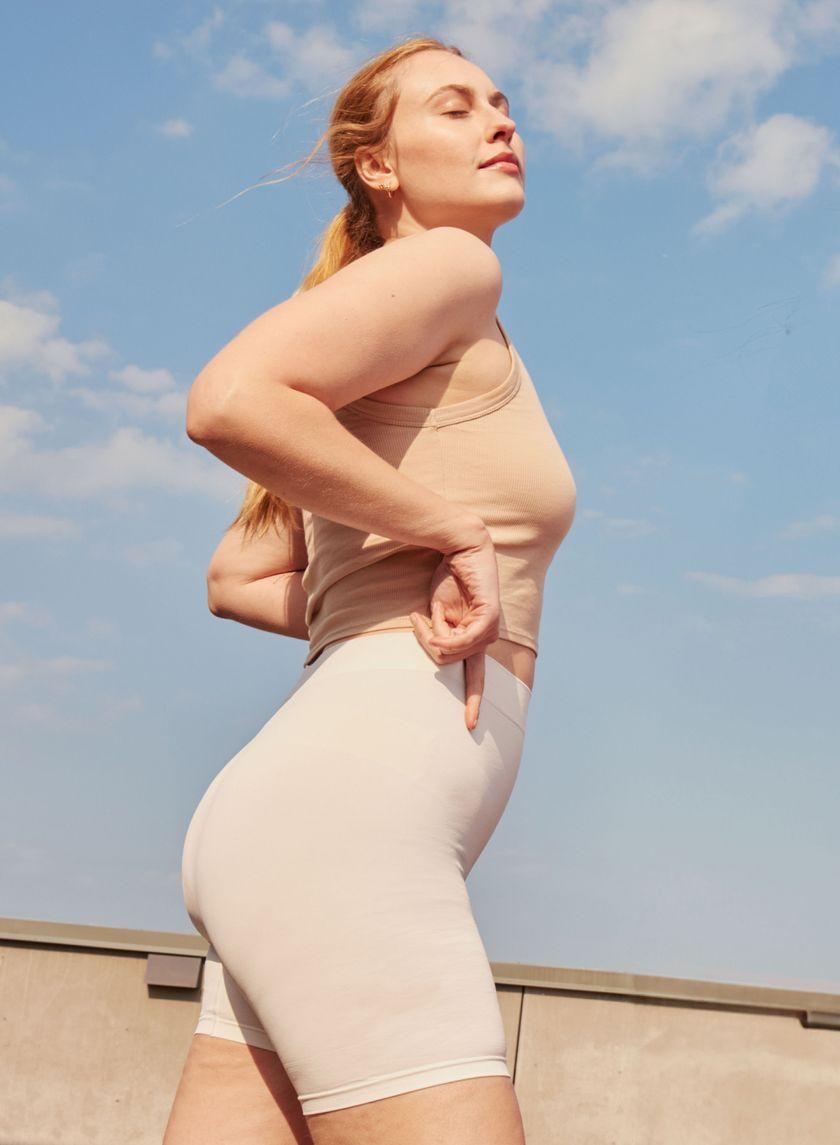 A blond woman looking up in the sky wearing a beige top and white slip shorts
